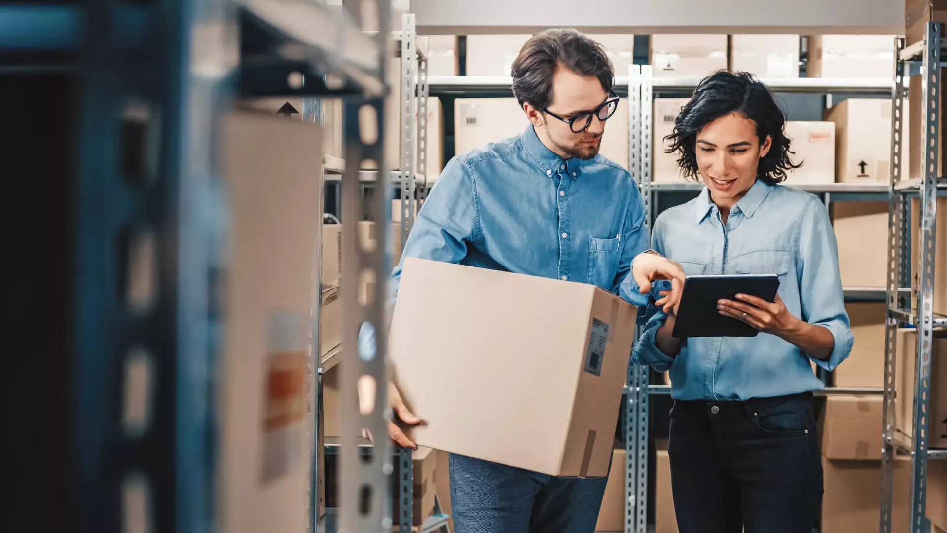 Female inventory manager shows digital tablet information to a worker holding cardboard box they talk and do work in the background stock of parcels with products ready for shipment