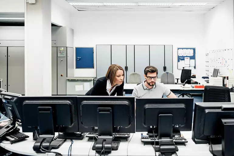 A printing factory operations manager assisting her colleague working at multiple computers in the control room